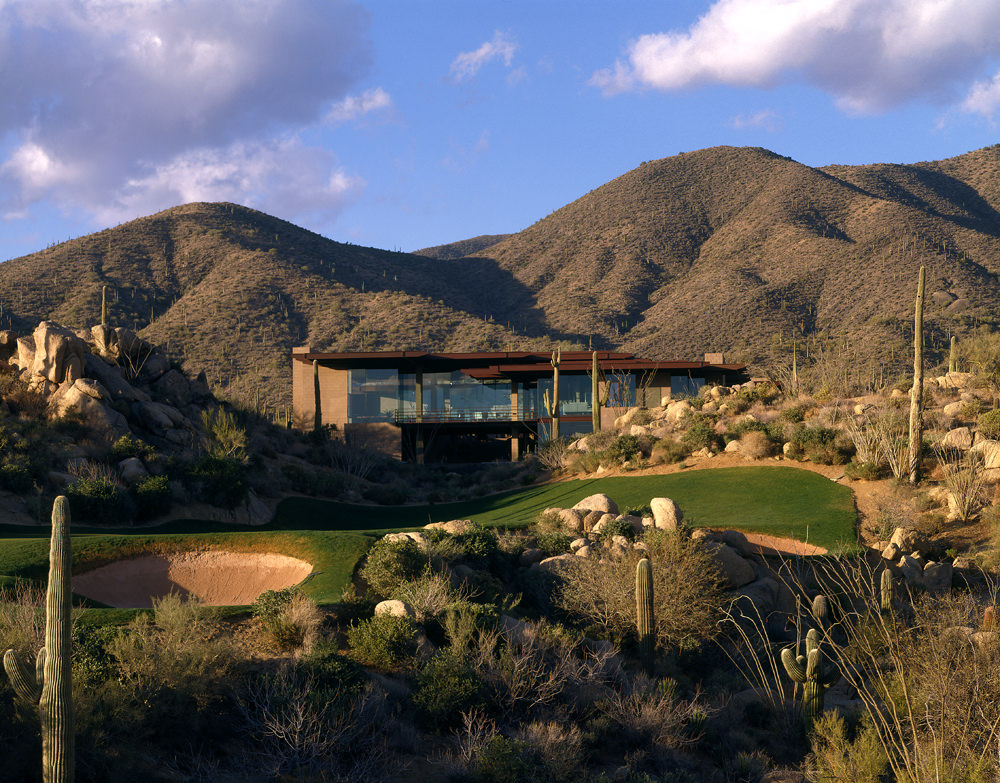 Exterior of Shadow Caster from afar, looking across a putting green at the home that is backdropped by desert mountains.