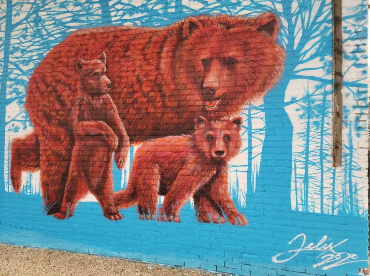 A vibrant mural painted on the side of a brick building depicts a brown bear mother with her two cubs in a forest of blue.