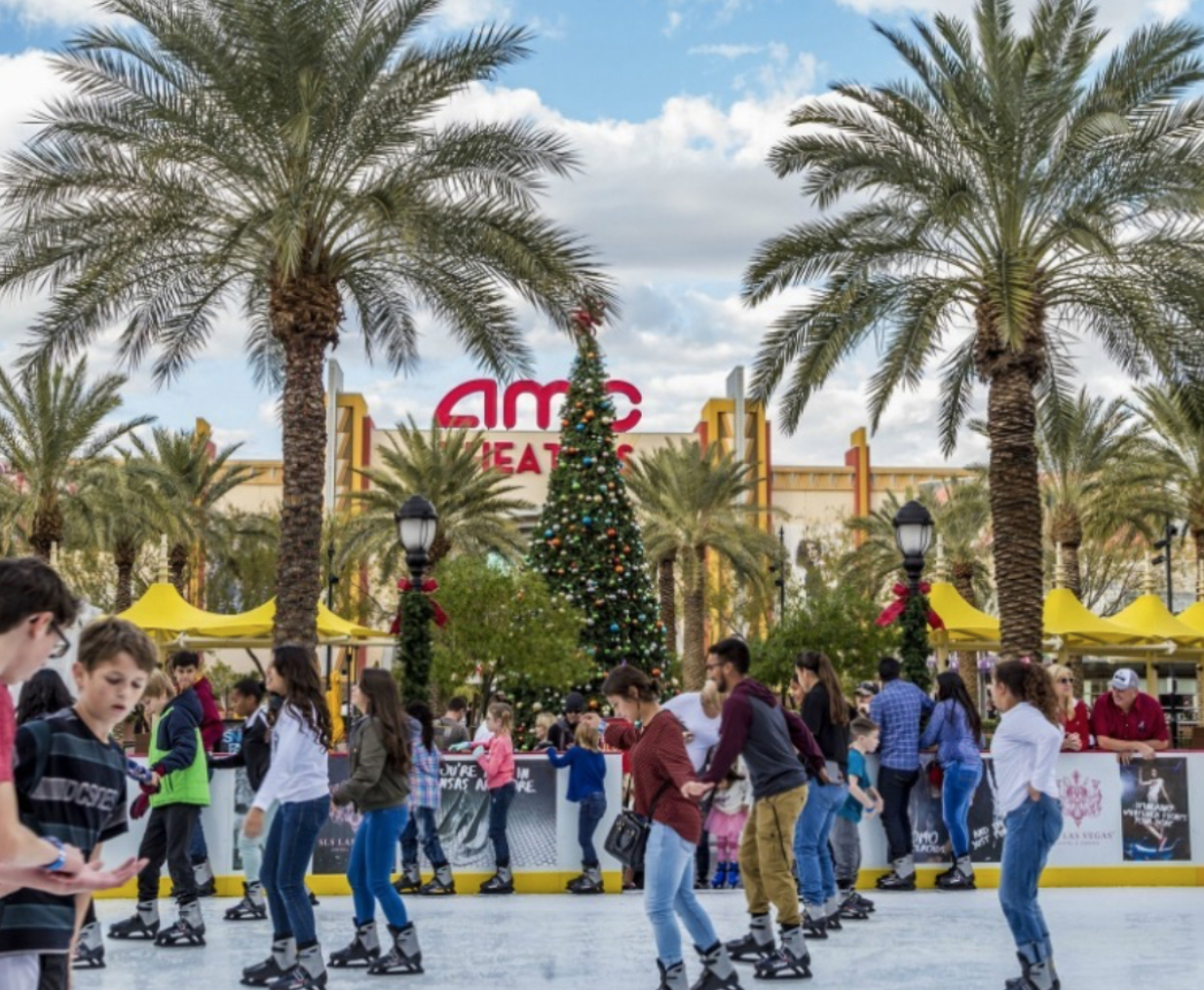 A crowd of people are iceskating in the foreground, in the background palm trees stand below an AMC Theater sign.